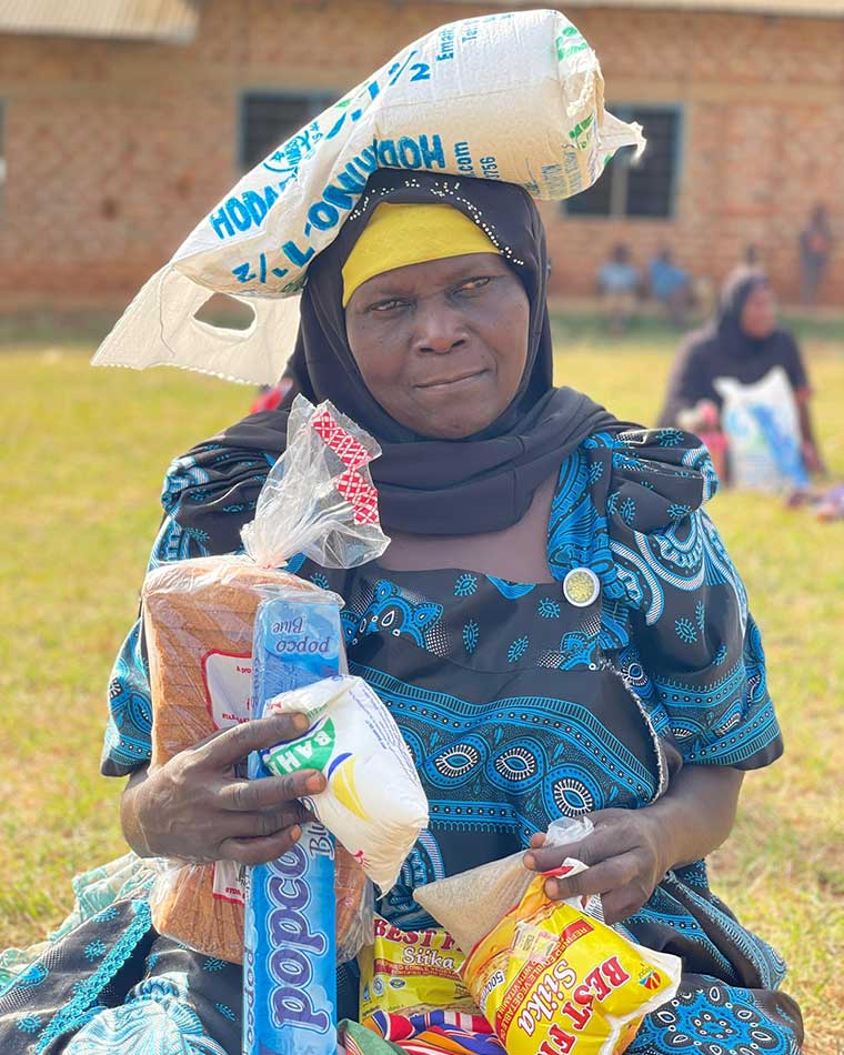 woman-wearing-gomesi-seated-on-ground-with-a-bag-of-maize-floor-balancing-on-the-head-after-recieving-food-aid-in-Eastern-Uganda